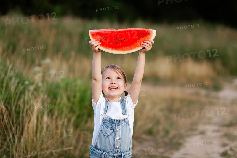 A cheerful child holds up a large watermelon slice against a lush summer field backdrop, capturing the essence of carefree childhood and joyful summer days