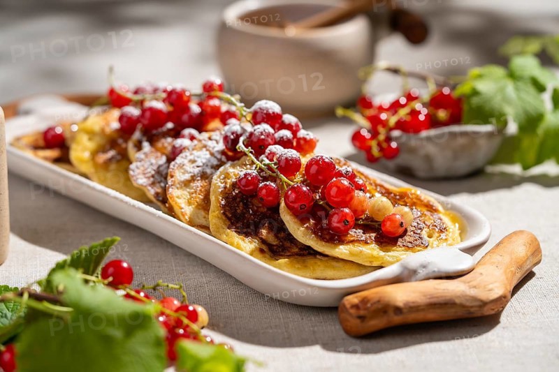Delicious pancakes adorned with red currants are served on a white ceramic plate. They rest on a textured beige fabric, creating a delightful breakfast scene