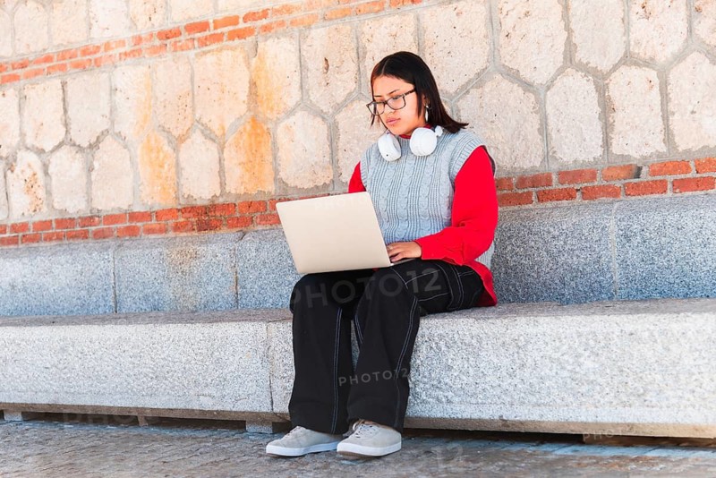 Hispanic student with glasses and headphones, focused on their laptop while seated on a stone bench outdoors Casual attire, including a red sweater and black jeans