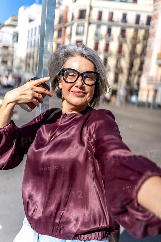 Mature woman happily capturing a moment by herself, holding a cup of coffee and wearing glasses, standing on a sunny city street, showing confidence and urban lifestyle