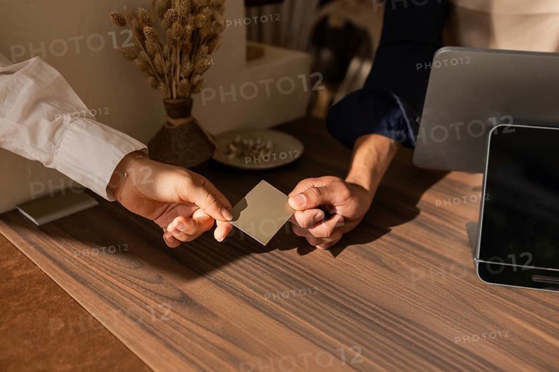 A customer and salesperson exchange a card over a stylish wooden counter in a cozy bag store, with a vase of dried flowers in the background adding a warm touch