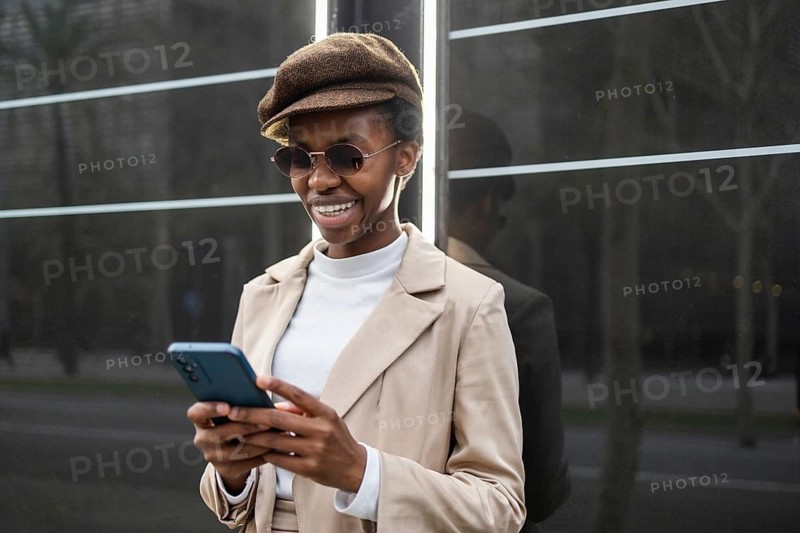 Urban black woman in a stylish outfit, wearing sunglasses and a cap, confidently uses her smartphone The modern city background adds to the chic, vibrant atmosphere