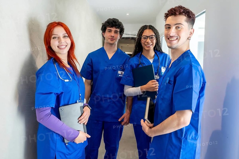 Diverse group of young medical students and healthcare professionals in blue scrubs smiling in a hospital corridor, holding a tablet and clipboard, symbolizing teamwork and training