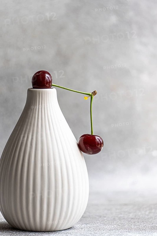 A ripe cherry delicately rests on an elegant white ceramic vase, with a soft concrete background adding texture to this minimalistic and stylish composition