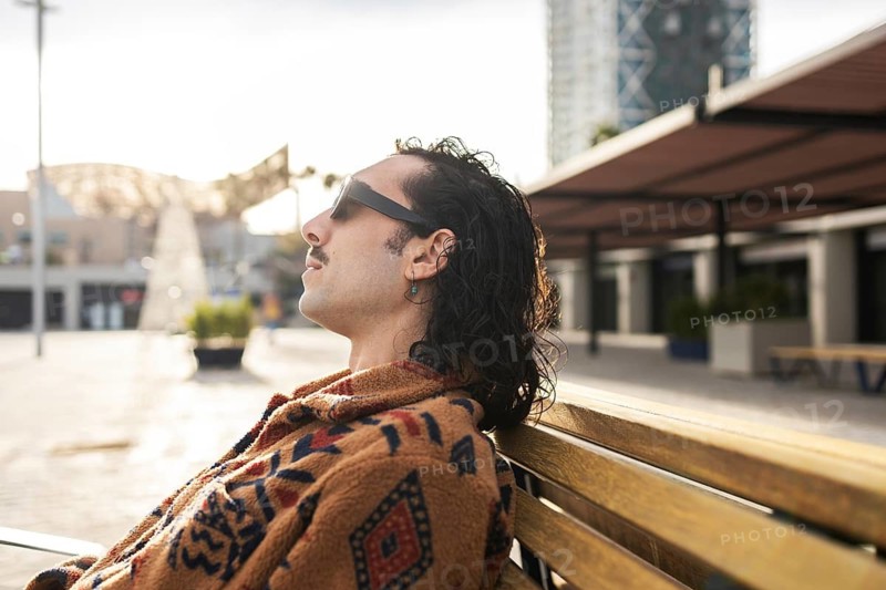 A man with long hair sits back on a wooden bench, wearing stylish sunglasses and a patterned jacket He seems relaxed, soaking up the sunshine in an urban setting