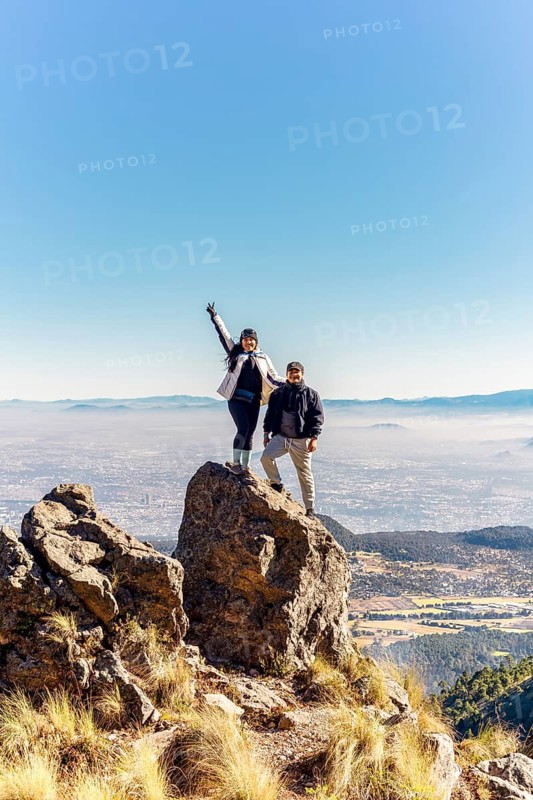 Two friends stand atop a rocky peak, enjoying a breathtaking mountain view under a clear blue sky Their joyous expressions capture the essence of adventure and freedom