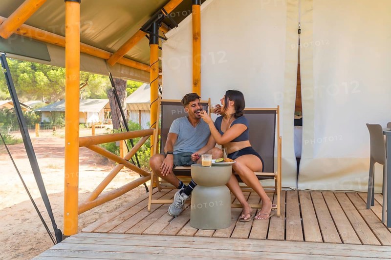 Young couple sharing breakfast and having fun on the wooden patio of their glamping tent, surrounded by nature and enjoying a relaxing vacation