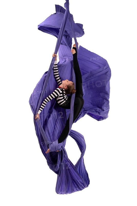 Full body side view of slim female aerial silks performer doing acrobatic splits while hanging on purple fabric against white background isolated