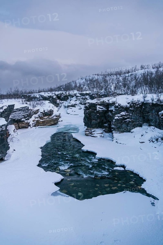 A striking view of a partially frozen river seamlessly cutting through the snow-covered terrain in Swedish Lapland, creating a stunning contrast between ice and water