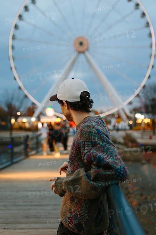 Side view of unrecognizable man wearing a cap and sweater, standing near a ferris wheel in Montreal, capturing the essence of urban fall vibes in Quebec