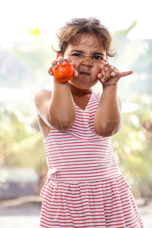 A child in a striped dress makes playful faces, holding a tangerine, enjoying a sunny day outdoors. The scene captures a moment of innocence and joy in summer
