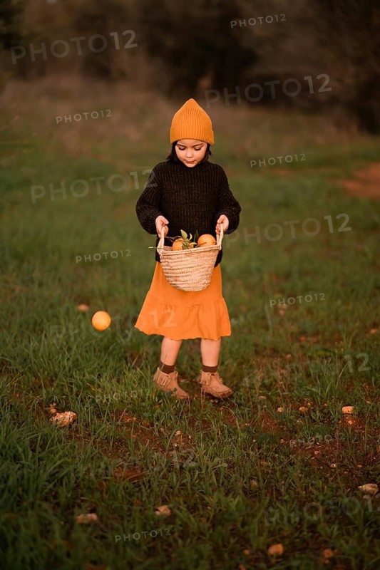 A child stands in a green field wearing a beanie, sweater, and skirt, holding a basket of oranges. The scene captures the essence of autumn in a rural setting