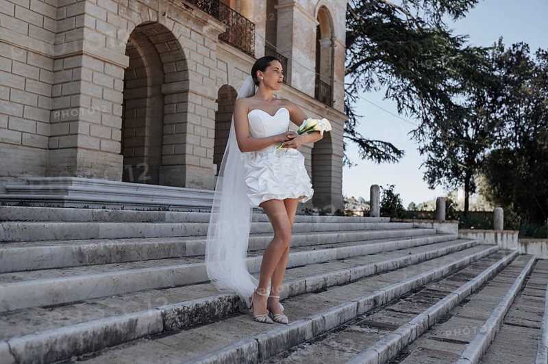 A bride in a short wedding dress holding a bouquet poses on stone steps in front of an ancient villa in Treia, province of Macerata, Italy, under a clear blue sky