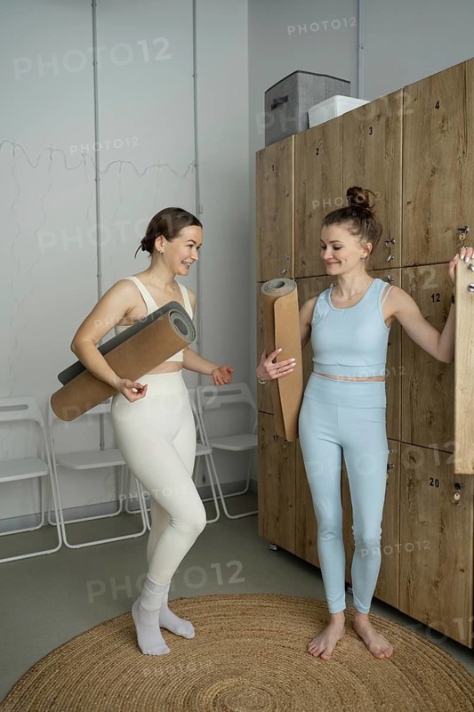 Two women in active wear chat animatedly in a modern locker room before a yoga class, holding mats and preparing for the session in a relaxed environment
