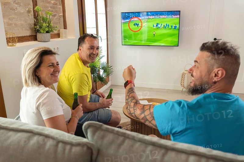 Friends celebrating a goal while watching a soccer match on TV at home. Smiling faces, raised arms, beer bottles and shared excitement in a cozy living room