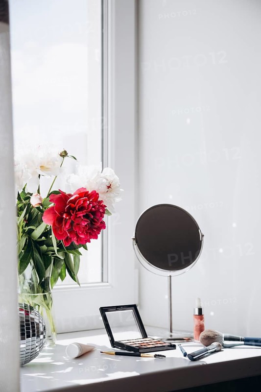 A vibrant bouquet of white and red peony flowers in a clear vase on a light-filled windowsill, accompanied by makeup items and a round mirror, presenting a fresh and elegant home interior scene