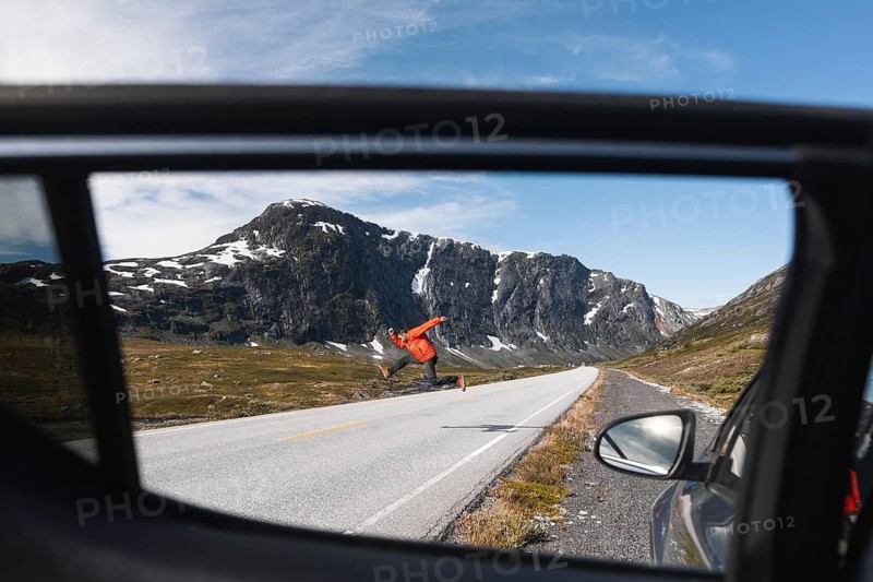 A traveler in a red jacket jumps joyfully on a mountain road in Norway, framed through a car window Snow-capped peaks and blue skies add to the charm of this adventure scene