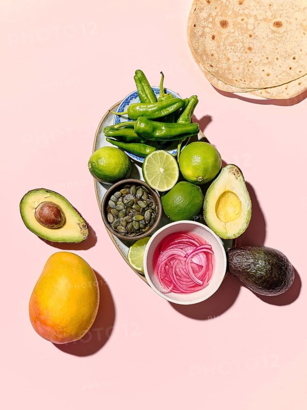 Top view of vibrant taco components spread on a pink background, featuring green peppers, limes, avocados, pumpkin seeds, pickled onions, and a ripe mango