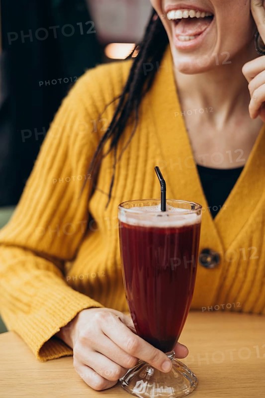 A woman in a cozy yellow sweater holds a tall glass of a coffee with a black straw, smiling joyfully The image captures a moment of refreshment and happiness