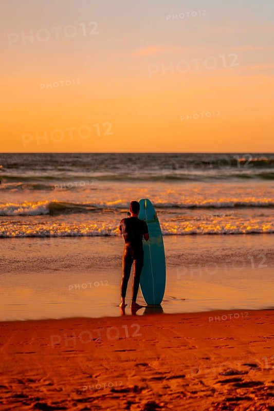 A man stands on the beach holding a blue surfboard. The sky is orange and the water is calm