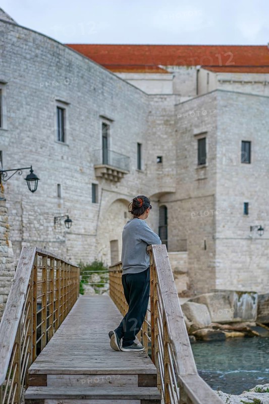 A visitor leans on a wooden bridge, gazing at the historic stone castle of Polignano a Mare, a charming coastal village on the picturesque Puglia coast