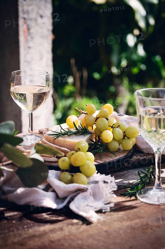 A serene outdoor still life composition featuring a glass of white wine and fresh grapes arranged on a rustic wooden table, basking in natural sunlight