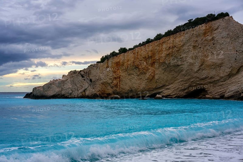Crystal-clear turquoise waters gently lap against the shores of Porto Katsiki beach in Lefkada, Greece, framed by dramatic cliffs and a cloudy sky