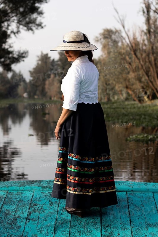 A woman in traditional clothing stands on a vibrant blue boat in Xochimilco, Mexico. The scenic waterway and lush greenery highlight the cultural richness of the region