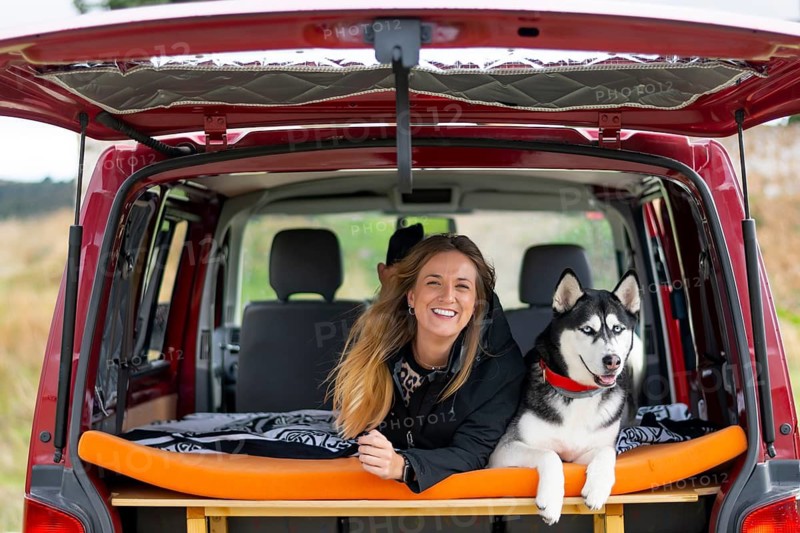 A joyful woman and her husky dog relaxing in the back of a red camper van, with a beautiful natural landscape visible, embodying the spirit of adventure and companionship