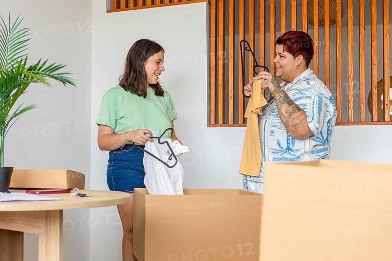 Lesbian friends happily unpack boxes in a new home, sorting clothes and sharing a moment of organization and camaraderie in moving house. The scene captures a fresh start and teamwork