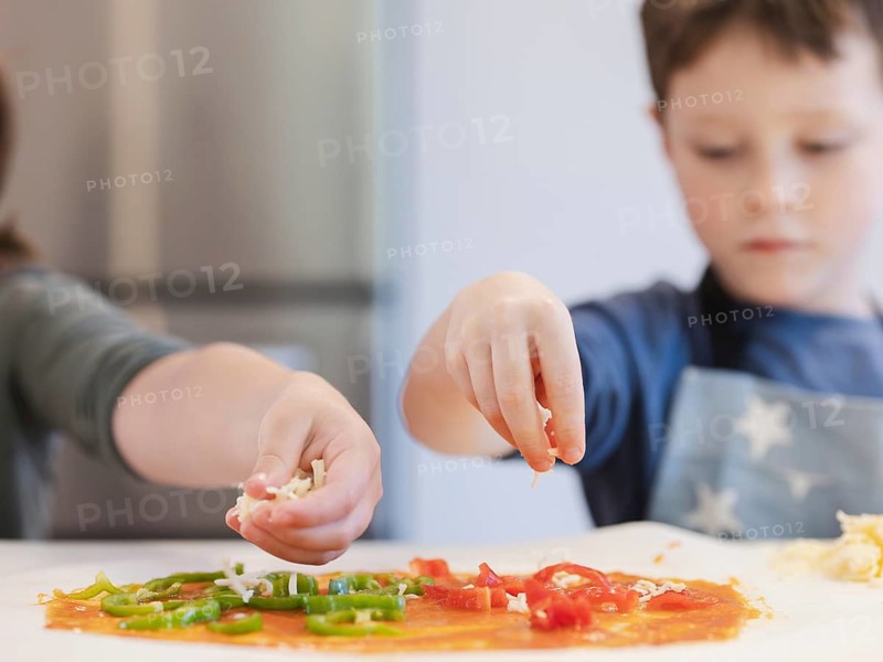 Close up of siblings hands sprinkling cheese and arranging vegetables on pizza dough. The scene captures creativity and teamwork in a fun cooking activity