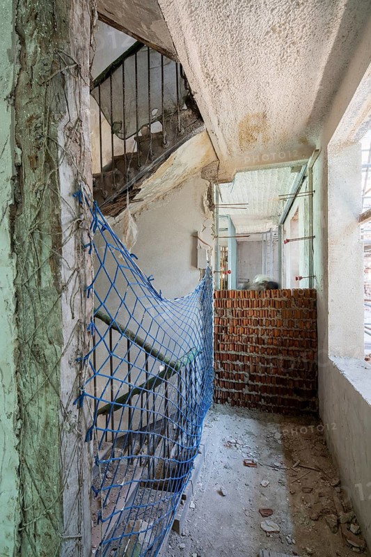 Construction site featuring a partially demolished wall, exposed brickwork, and a blue safety net. The staircase area shows ongoing renovation activities in a building