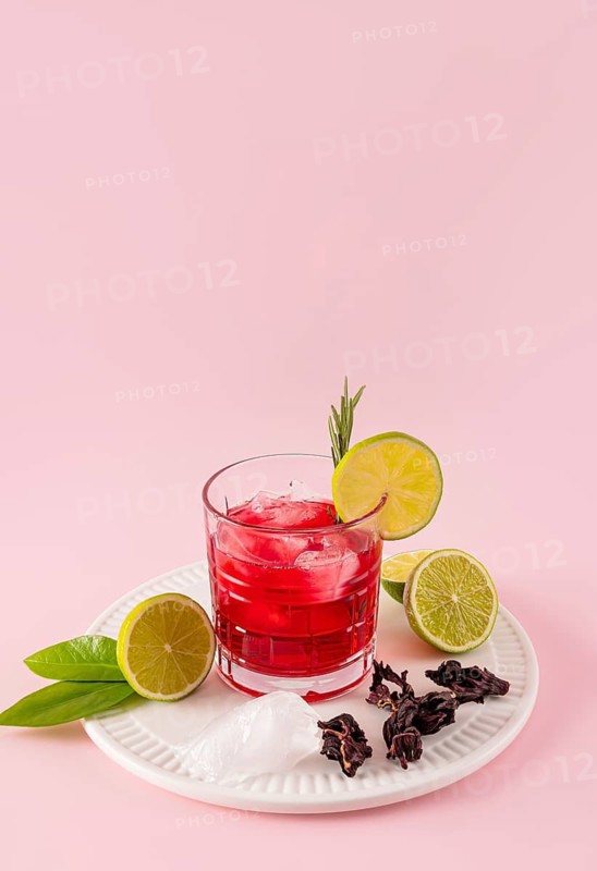 Vibrant hibiscus tea with ice and lime slices on a white plate. Enhanced with hibiscus flowers and green leaves, set against a soft pink background, offering a refreshing appeal