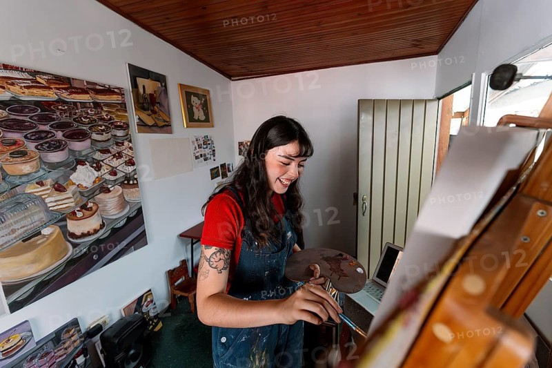 A young Latin woman engrossed in painting on a canvas in her home studio, surrounded by vivid artwork. She holds a palette and paintbrush, expressing creativity and passion
