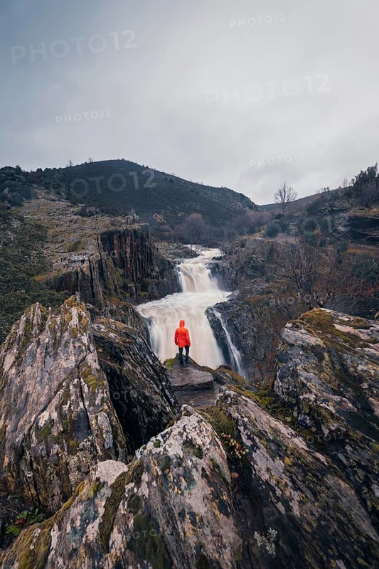 Back view of an unrecognizable person in a bright red jacket admiring the majestic Cascada del Aljibe waterfall amidst rocky terrain under an overcast sky in Guadalajara, Spain