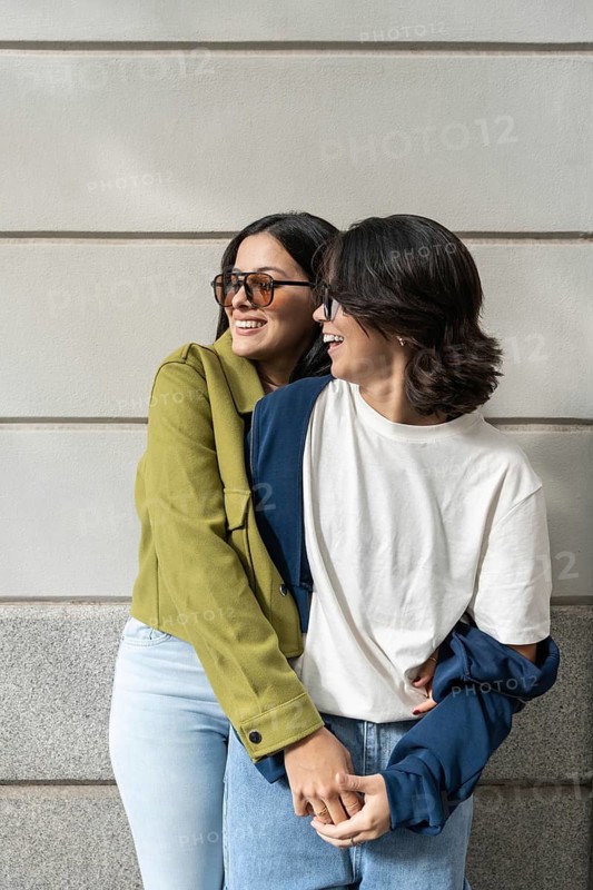 A joyful lesbian couple embraces outdoors. Wearing casual outfits and sunglasses, they smile warmly while sharing a tender moment, symbolizing love and happiness in everyday life