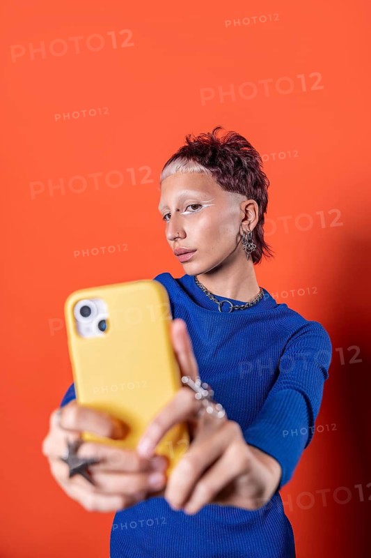 A stylish woman takes a selfie against a vivid orange backdrop, wearing a blue sweater and holding a yellow smartphone. Her bold makeup and unique accessories add flair