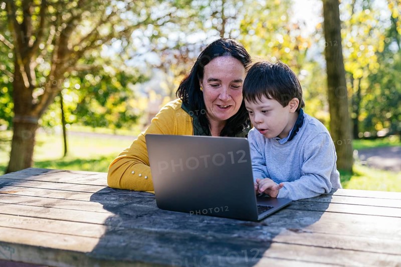A mother and her son with Down syndrome engage in learning with a laptop while sitting at a wooden table in a sunny park. The greenery around creates a peaceful atmosphere