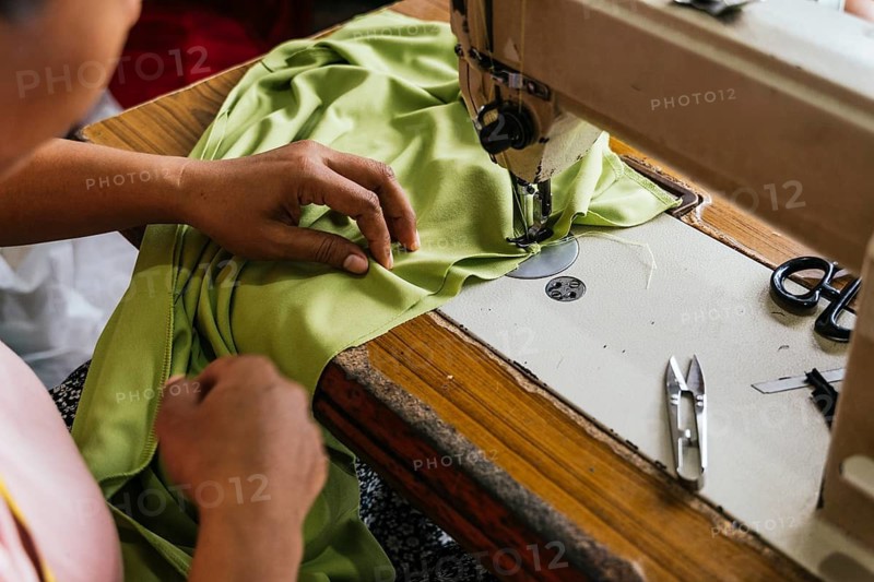 Close up of asian seamstress hands guiding green fabric through a sewing machine. Essential tools like scissors and a seam ripper are nearby, highlighting the asian seamstress workspace
