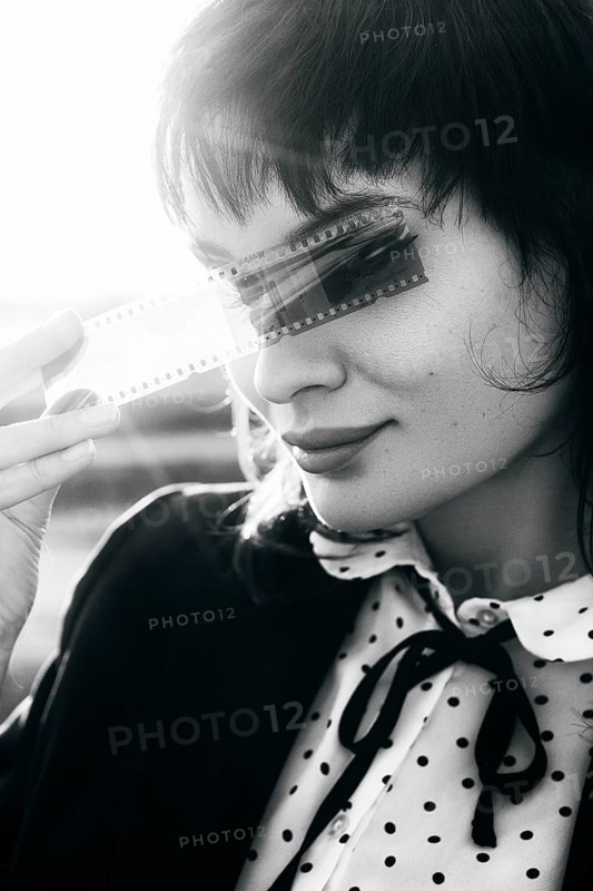 Black and white portrait of a woman holding a film strip over her eyes, evoking a sense of mystery and nostalgia, with sunlight in the background