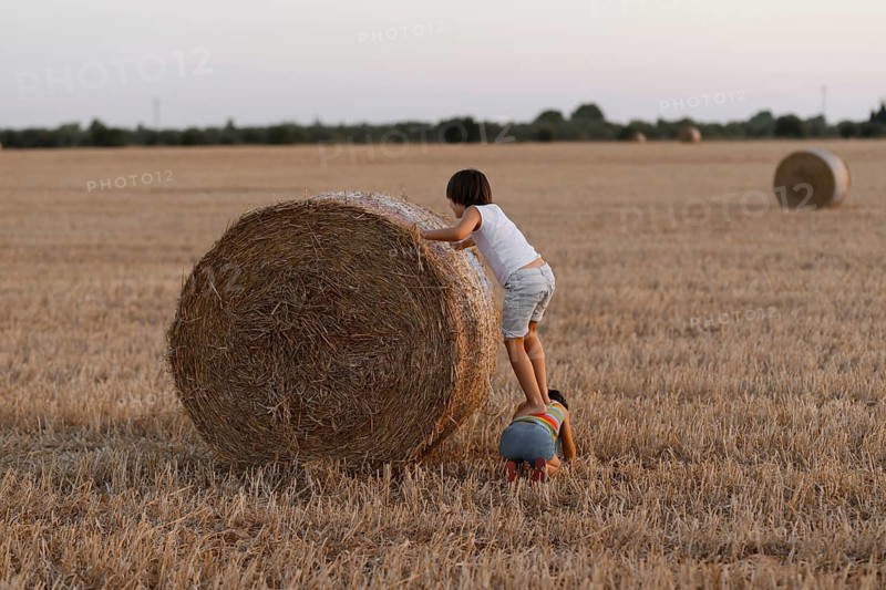 Two siblings interact with a large hay bale in a field, displaying curiosity and playfulness amidst an agricultural setting under a dusky sky