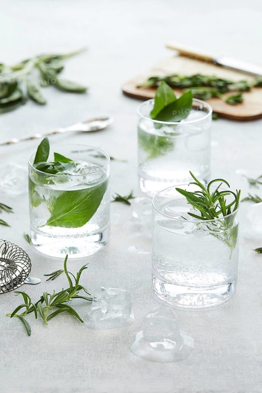 Refreshing beverages in glasses, garnished with green rosemary and mint, accompanied by scattered herbs and ice on a textured surface