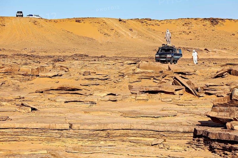 An off-road vehicle and people standing in front of sand dunes in a vast, rocky desert landscape, By off-road vehicle through the Sahara desert in Algeria