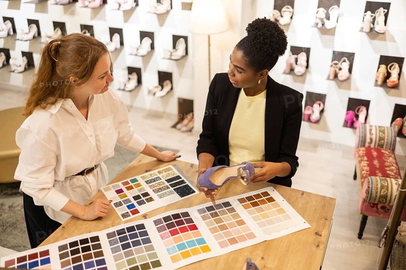 Two women collaborate over color swatches and shoe selection in a bridal studio environment, focusing on coordinated shoe designs and color matching