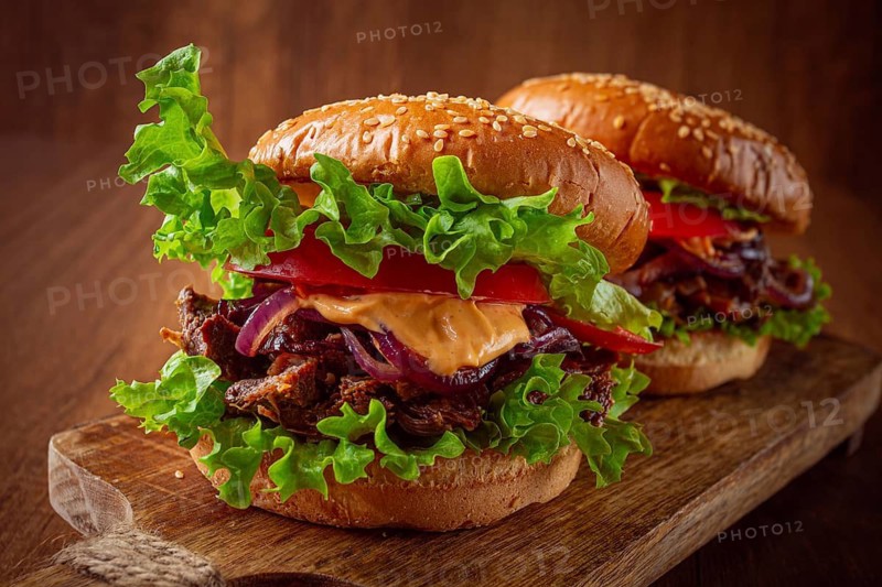 Two burgers are stacked high with fresh lettuce, tomato slices, grilled onions, and a savory sauce. They sit on a wooden board in a warm light setting. Ideal for a meal or snack