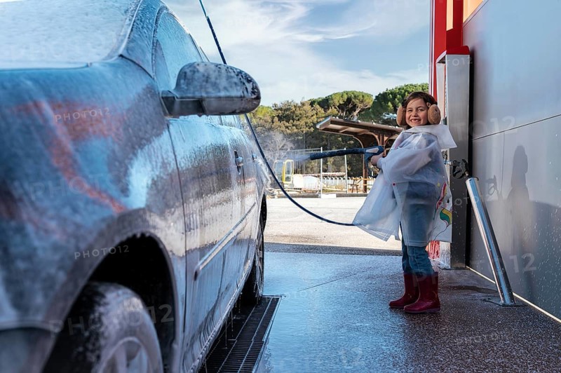 A joyful child in a clear raincoat and earmuffs sprays water on a foam covered car at a self serve car wash, enjoying the outdoor activity on a sunny day