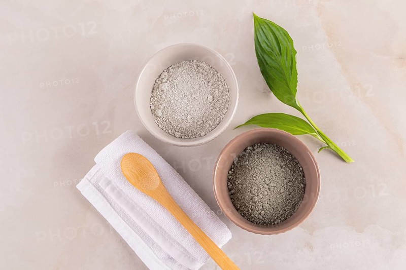 Top view of two bowls of cosmetic clay powder, a wooden spoon, and fresh leaves on a marbled background, ideal for skincare and beauty routines