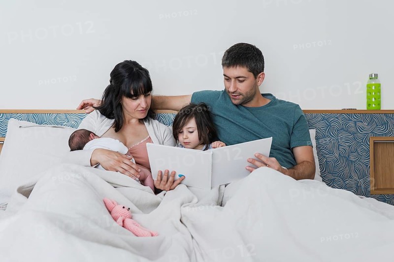 A loving young family shares an intimate moment, with parents and their children, including a big sister and a newborn baby, enjoying storytime in bed together