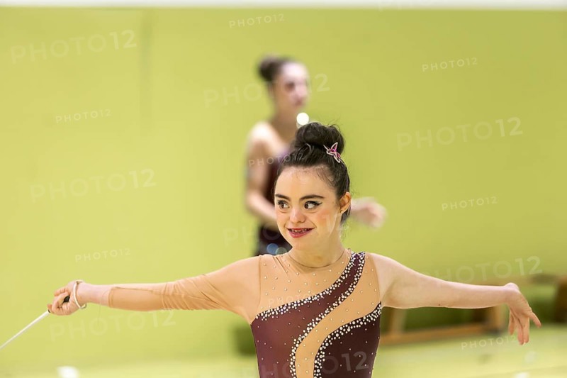 A disabled rhythmic gymnast with a cheerful smile and focus, wearing a sparkly costume, practices with a ribbon in a bright gym setting, emphasizing joy and dedication to the inclusive sport