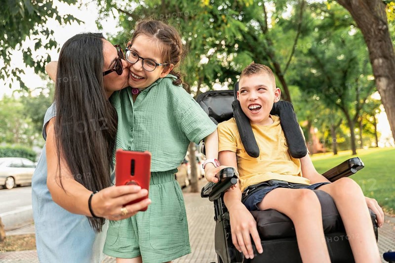 A joyful family enjoys a sunny day outdoors, capturing a selfie. The mother kisses her daughter with tracheostomy, while her son with cerebral palsy smiles in his wheelchair